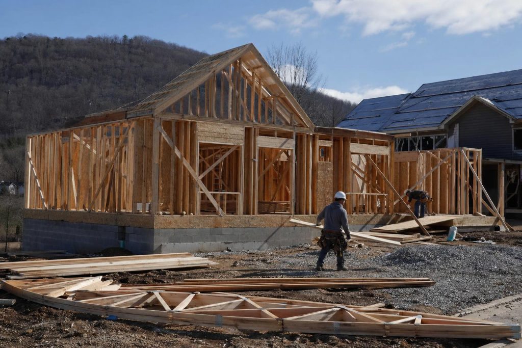A construction worker stands in front of a residential home during the framing stage, with exposed wooden beams and mountain backdrop visible in daylight.