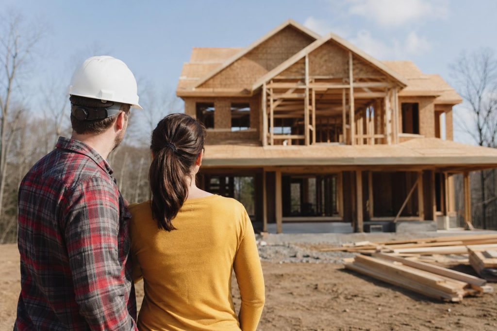A man and woman in hard hats and work clothing inspect a two-story residential house frame under construction on a clear day.